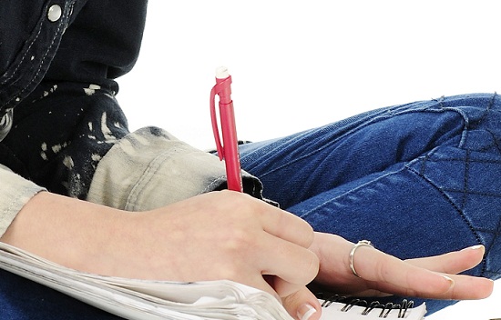 An attractive young teen happily sitting cross-legged on the floor while writing in her note pad. On a white background.